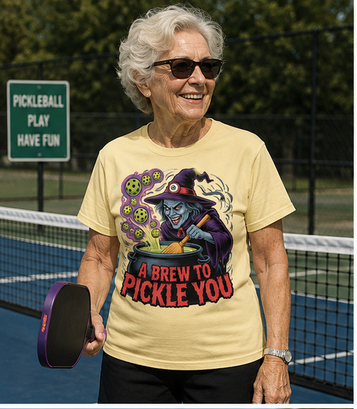 An older woman wearing sunglasses and a pale yellow graphic T-shirt stands on a pickleball court holding a paddle. The shirt features a witch stirring a cauldron with the text 'A BREW TO PICKLE YOU.