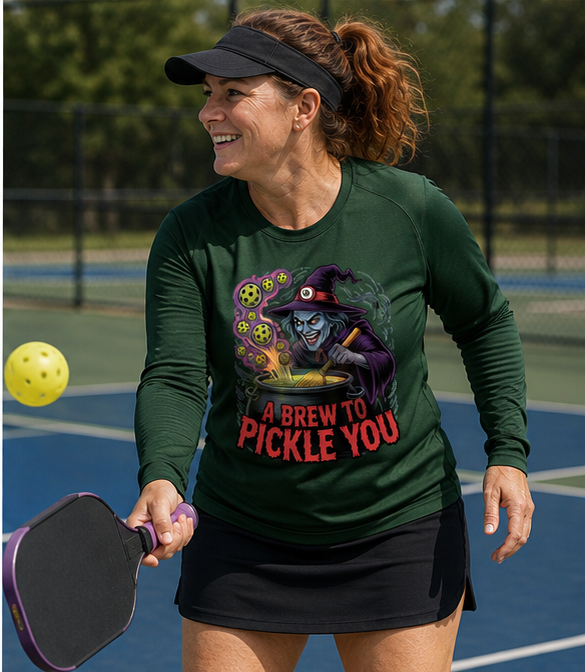 A woman playing pickleball on an outdoor court wearing a dark green long-sleeve graphic shirt with a witch illustration and the text 'A BREW TO PICKLE YOU'.