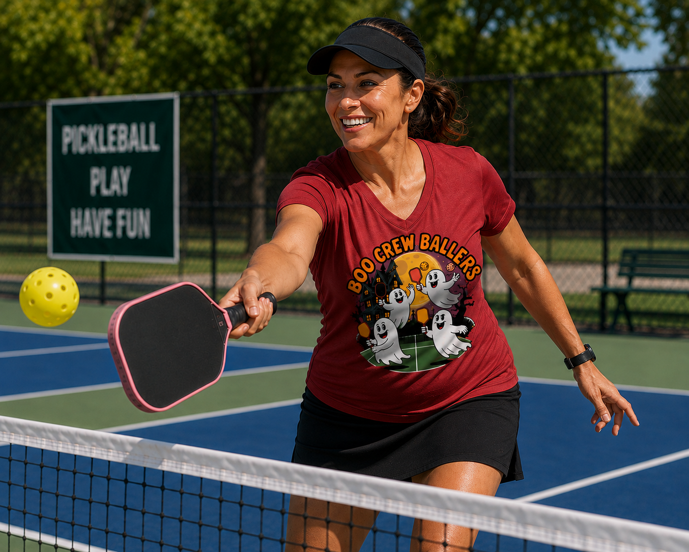A woman playing pickleball on an outdoor court, wearing a red graphic t-shirt that reads 'BOO CREW BALLERS' with cartoon ghosts, a black visor, and a black skirt.