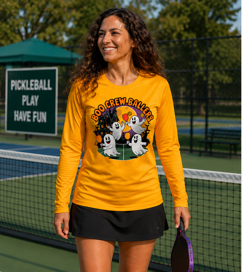 A woman stands on a pickleball court wearing a bright yellow long-sleeve graphic shirt with a Halloween ghost design and the text 'BOO CREW Ballers,' paired with a black skirt. A pickleball sign and court are visible in the background.