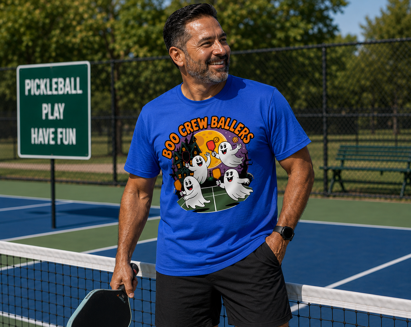 A man wearing a bright blue graphic t-shirt with 'BOO CREW BALLERS' and cartoon ghosts printed on the front, standing on a pickleball court holding a paddle.