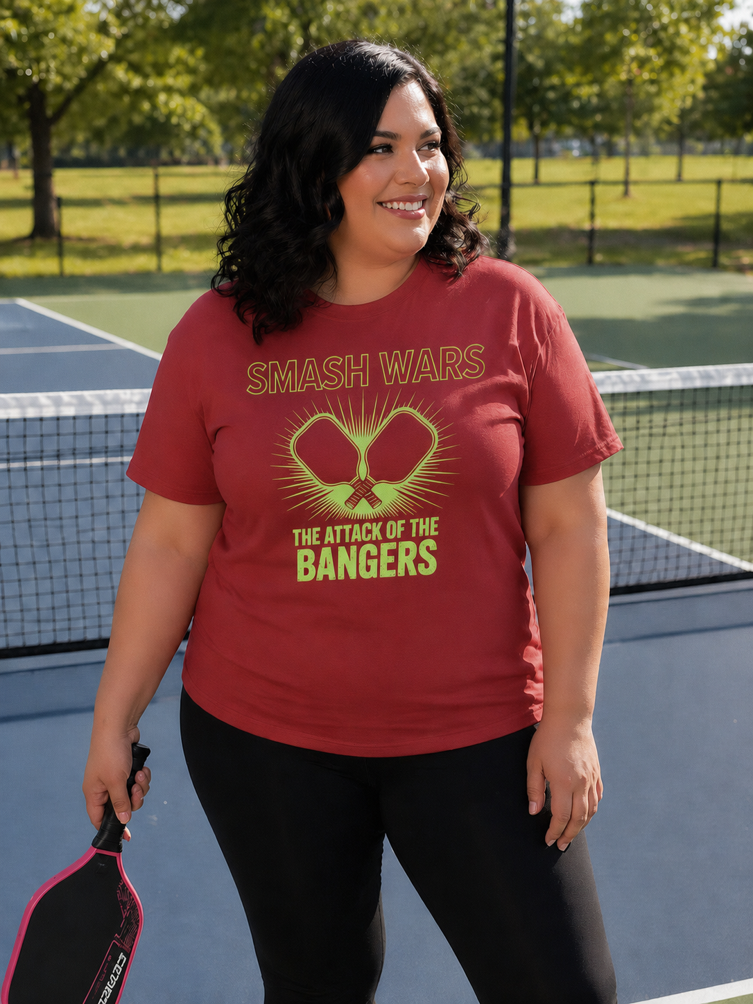 "Woman holding a pickleball paddle on an outdoor court wearing the Smash Wars Attack of the Bangers graphic tee in red"