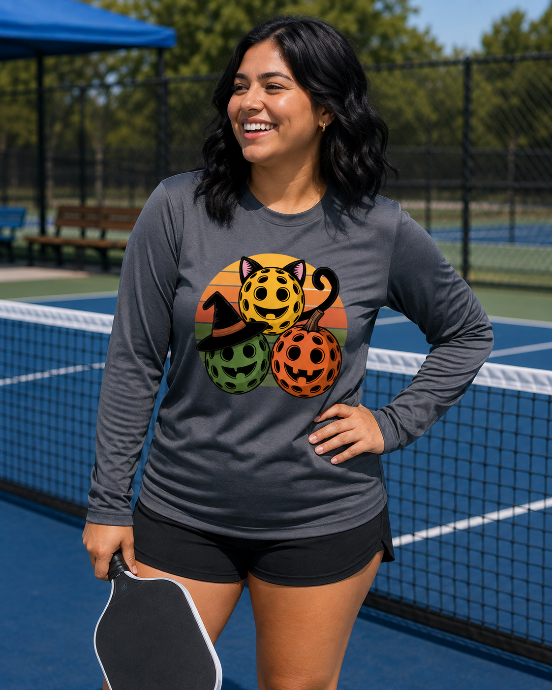 A woman wearing a dark gray long-sleeve athletic shirt with a colorful graphic of three Halloween pickleballs, a witch-pumpkin-cat hat, standing on a blue pickleball court holding a paddle.