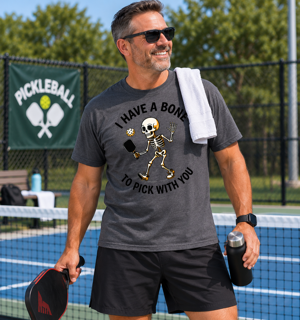 Man wearing a charcoal gray “I Have a Bone to Pick with You” pickleball T-shirt while holding a paddle and water bottle on a pickleball court.⠋