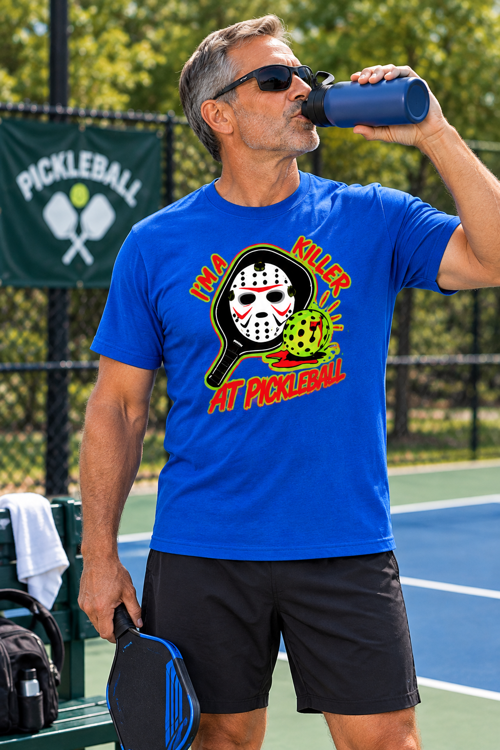 Man wearing a blue “I’m a Killer at Pickleball” T-shirt while drinking from a water bottle and holding a paddle on a pickleball court.