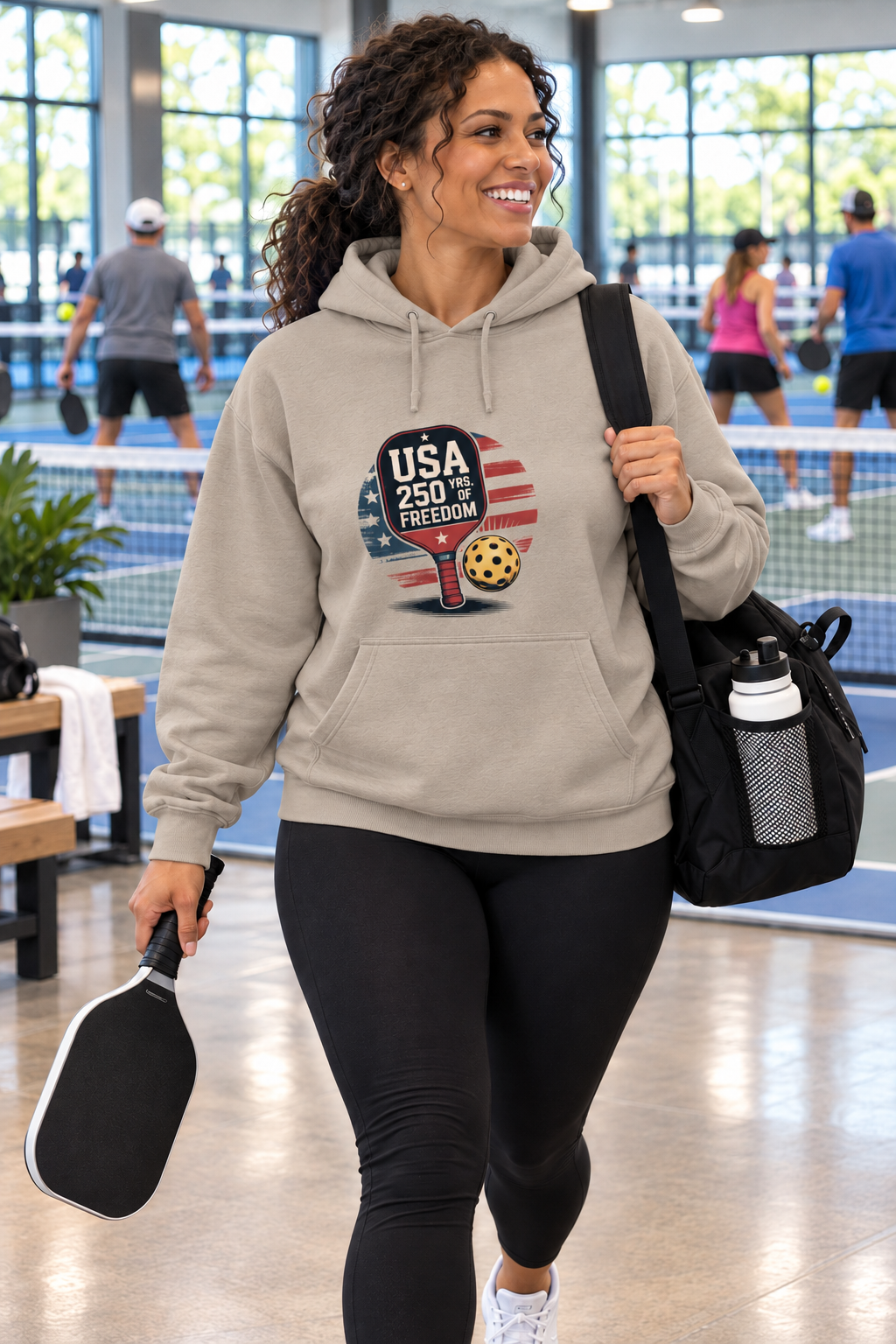 Woman wearing a sand hoodie with a patriotic pickleball graphic reading “USA 250 Yrs. of Freedom,” carrying a paddle and gear bag at an indoor pickleball court.