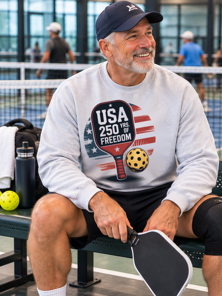 Man seated beside an indoor pickleball court holding a paddle, wearing a light gray “USA 250 Yrs. of Freedom” sweatshirt with an American flag, paddle, and ball graphic.