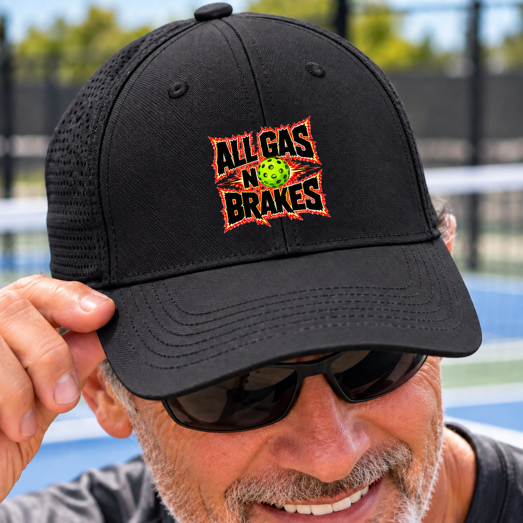 Smiling man in sunglasses wearing a black mesh-back cap with an “All Gas No Brakes” design and a green pickleball, photographed beside a pickleball court.⠋