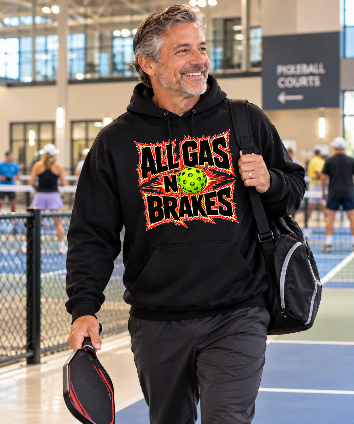 A smiling man walking on an indoor pickleball court wearing a black hoodie with a colorful 'ALL GAS NO BRAKES' graphic on the chest, holding a paddle and carrying a black bag.