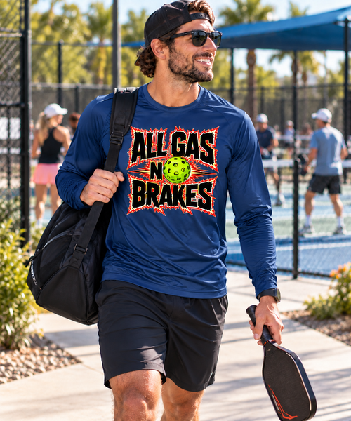 Smiling man in sunglasses and a backwards cap walks past sunny outdoor pickleball courts carrying a paddle and a black gear bag, wearing a navy long-sleeve shirt with a bold "All Gas No Brakes" graphic and a neon green pickleball in the center.