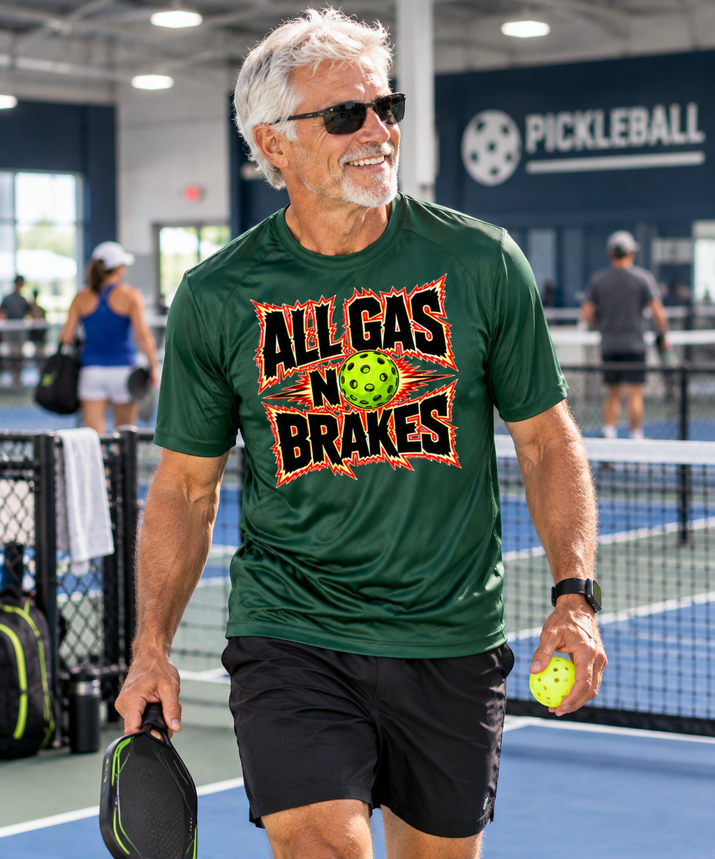 Man in sunglasses walking on an indoor pickleball court, holding a paddle and ball while wearing a dark green T-shirt with an “All Gas No Brakes” pickleball graphic.