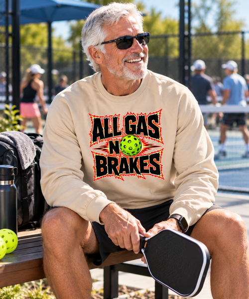 A smiling older man wearing sunglasses sits on a bench at a pickleball court, wearing a beige crewneck sweatshirt with a graphic that reads 'ALL GAS NO BRAKES' in black and red with a green pickleball in the center. He holds a black paddle, with a water bottle and pickleballs on a bench beside him.