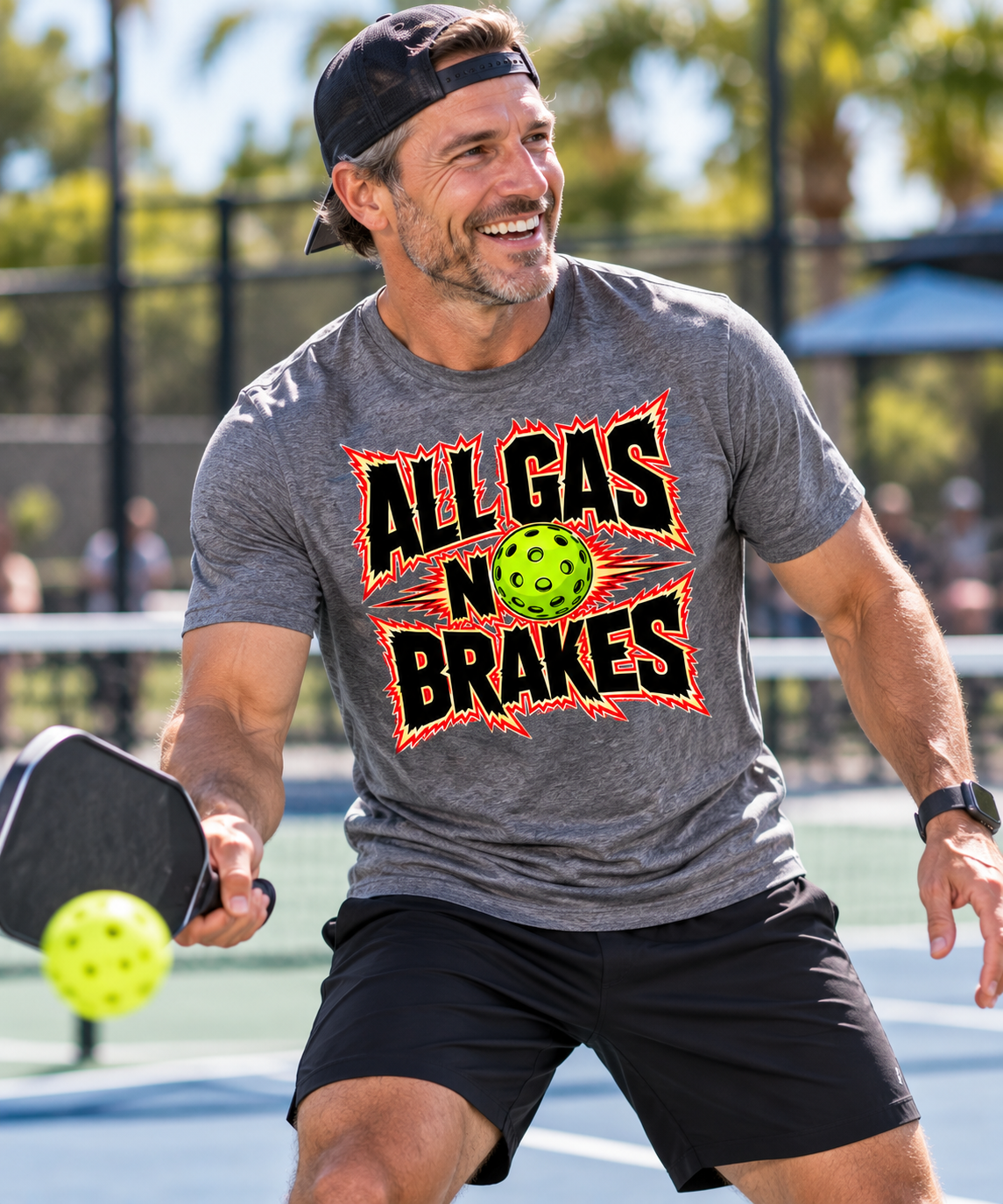 A smiling man playing pickleball wears a gray “All Gas No Brakes” T-shirt with a pickleball graphic.