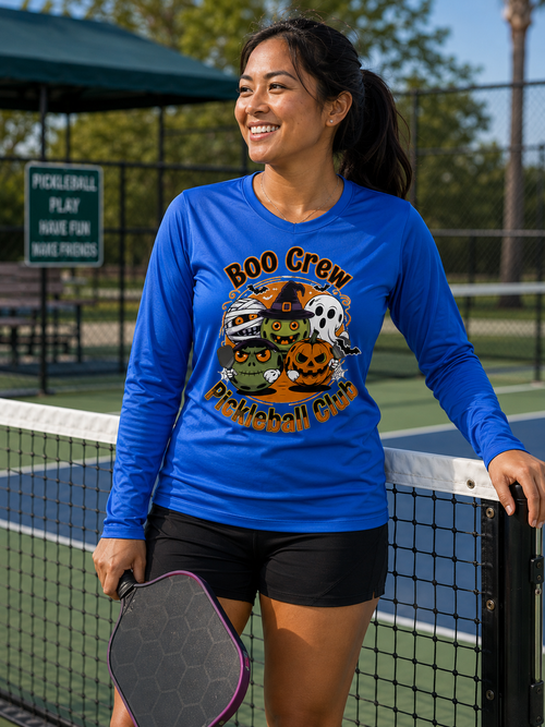 A woman wearing a bright blue long-sleeve graphic shirt stands on a pickleball court, holding a pickleball paddle. The shirt features a colorful Halloween-themed graphic that reads 'Boo Crew Pickleball Club' with pumpkins, a ghost, a mummy, and a witch.