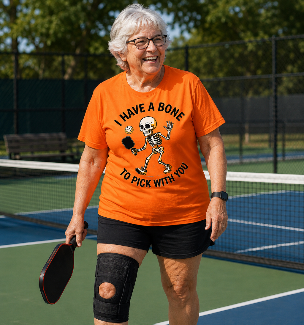 An older woman on a pickleball court wearing a bright orange graphic T-shirt that reads 'I HAVE A BONE TO PICK WITH YOU' with a cartoon skeleton, black shorts, and a knee brace, holding a paddle.