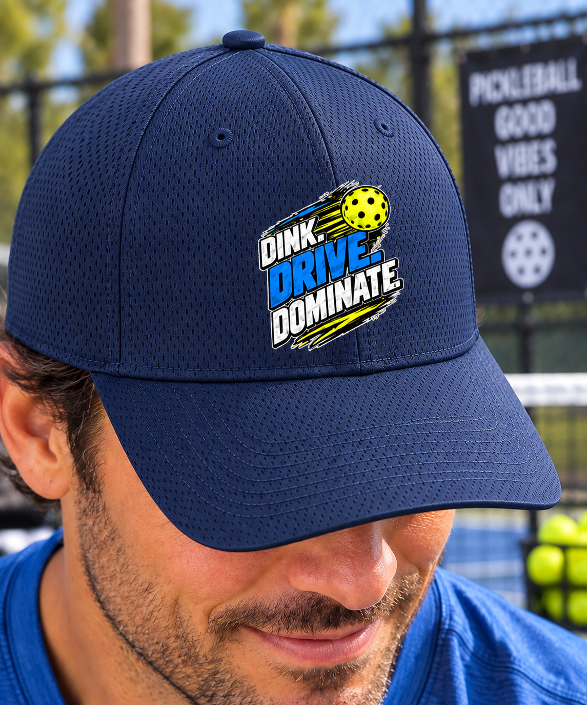 A man wearing a navy blue mesh baseball cap with a custom embroidered graphic that reads 'DINK DRIVE DOMINATE' in blue, white, and yellow, with a pickleball motif. The background shows a blurred pickleball court and net.