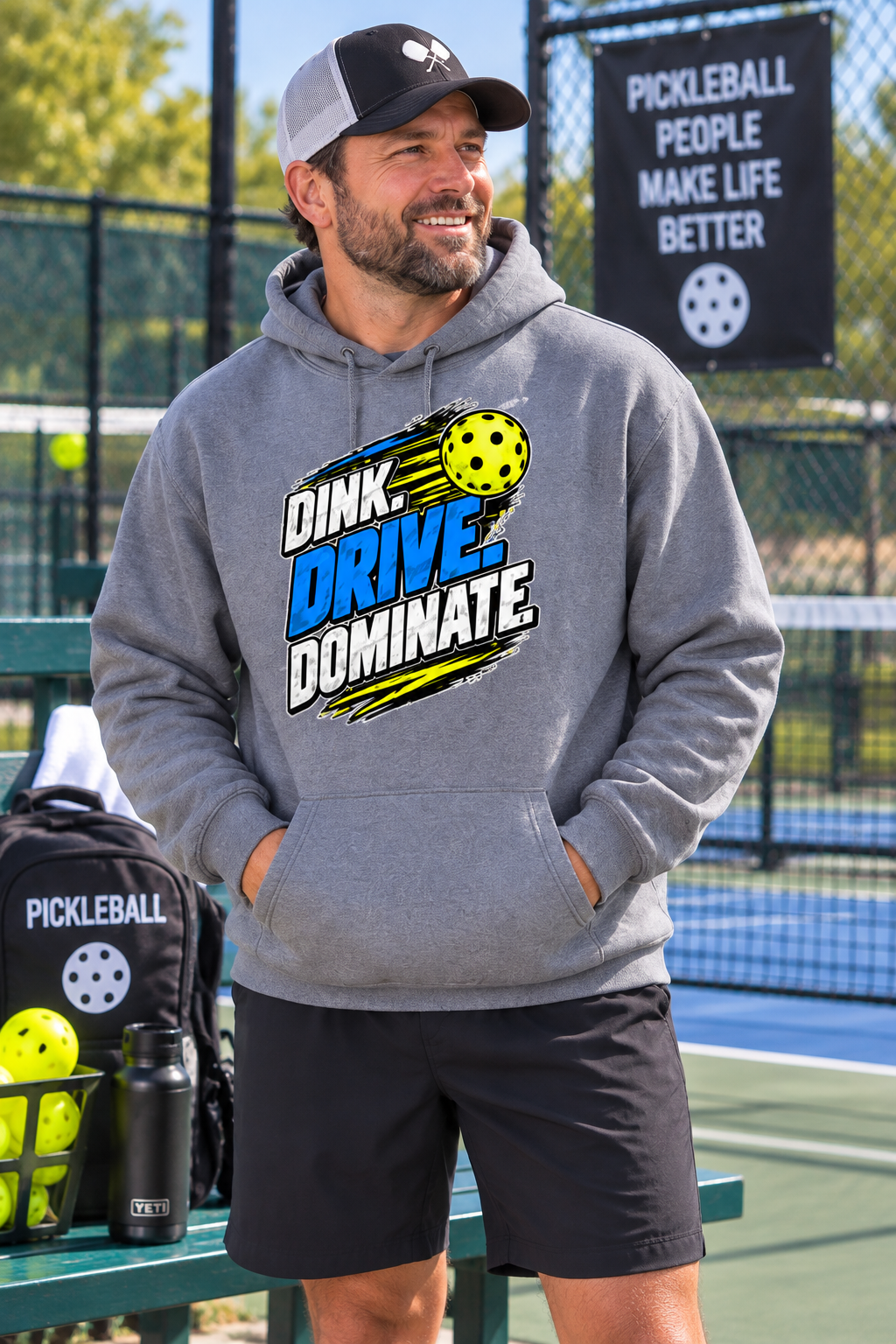 A man wearing a gray hoodie with a 'DINK DRIVE DOMINATE' pickleball graphic stands on an outdoor pickleball court. The hoodie features a bold graphic with yellow, blue, and white text and a pickleball illustration.