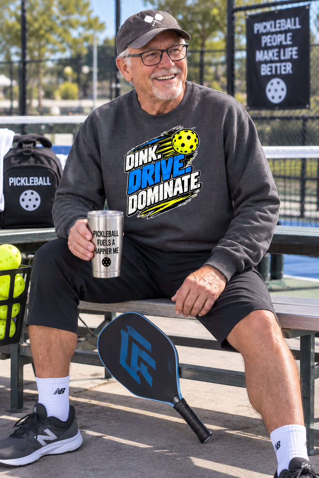An older man sitting on a bench at a pickleball court wearing a dark gray sweatshirt with a 'DINK DRIVE DOMINATE.' graphic, black shorts, and holding a pickleball mug.
