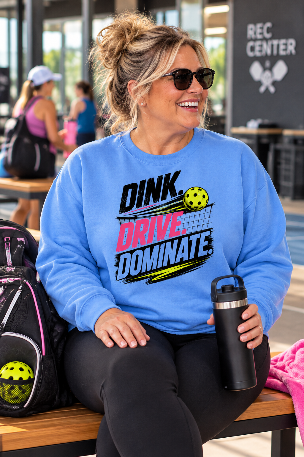 Woman wearing a blue “Dink Drive Dominate” pickleball sweatshirt, seated on a recreation center bench with a water bottle and pickleball bag.