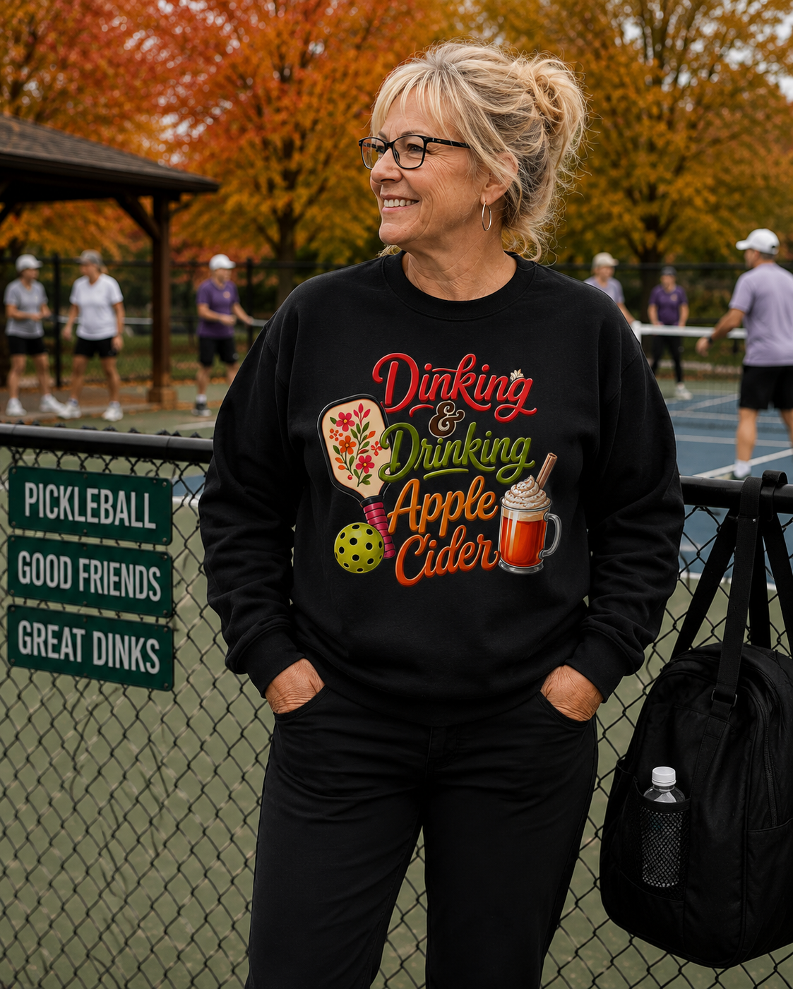 "Older woman smiling and wearing a black 'Dinking & Drinking Apple Cider' graphic crewneck sweatshirt and glasses while leaning against a fence with players rallying in the background, a 'Pickleball Good Friends Great Dinks' sign, and autumn foliage"