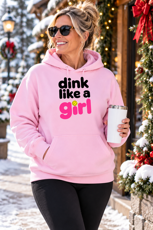 Woman wearing a pink “Dink Like a Girl” pickleball hoodie and sunglasses, holding a white tumbler on a snowy, holiday-decorated street.