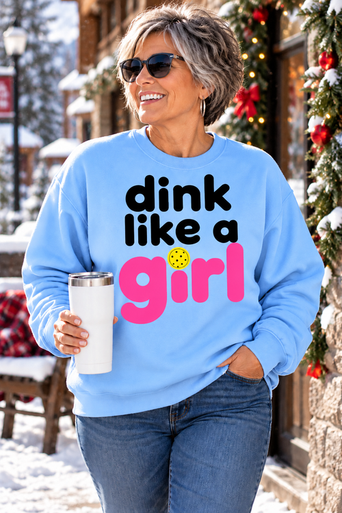 Woman wearing a light blue “dink like a girl” sweatshirt with black and pink lettering and a yellow pickleball graphic, holding a white tumbler on a snowy, holiday-decorated street.