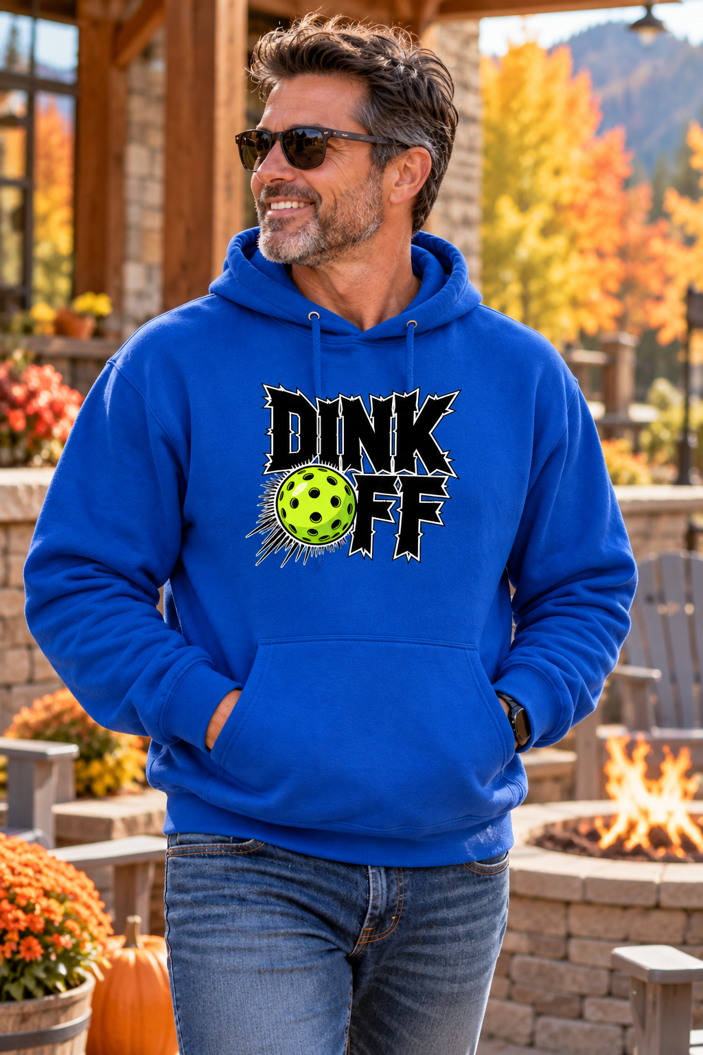 Man wearing a royal blue “Dink Off” pickleball hoodie with a neon green ball graphic, standing on an autumn patio beside a fire pit.