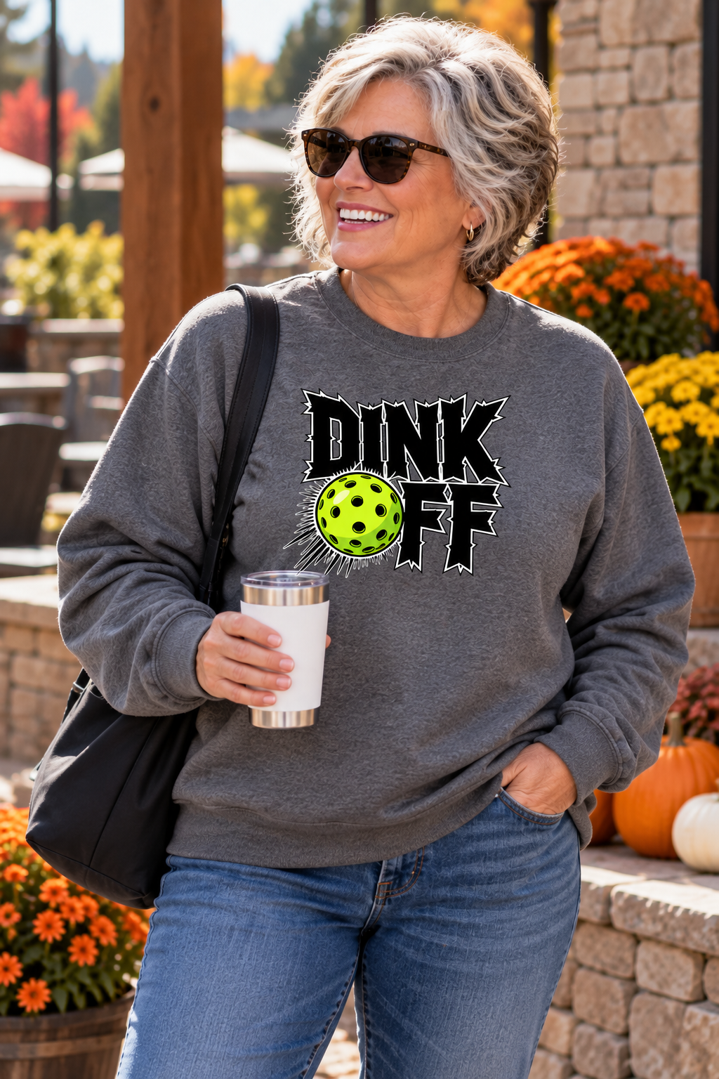 Woman wearing a gray “DINK OFF” pickleball sweatshirt with a neon green ball graphic, holding a tumbler outdoors among fall flowers and pumpkins.