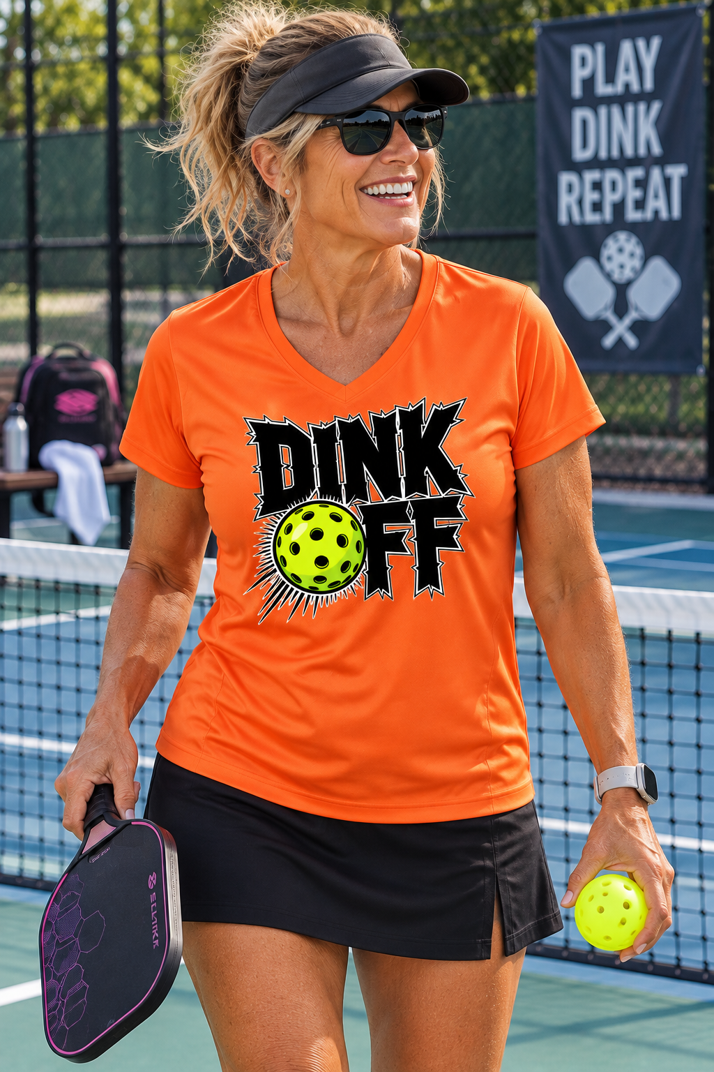 Woman wearing an orange “Dink Off” pickleball V-Neck moisture wicking T-shirt with a neon yellow pickleball graphic, holding a paddle and ball on an outdoor pickleball court.