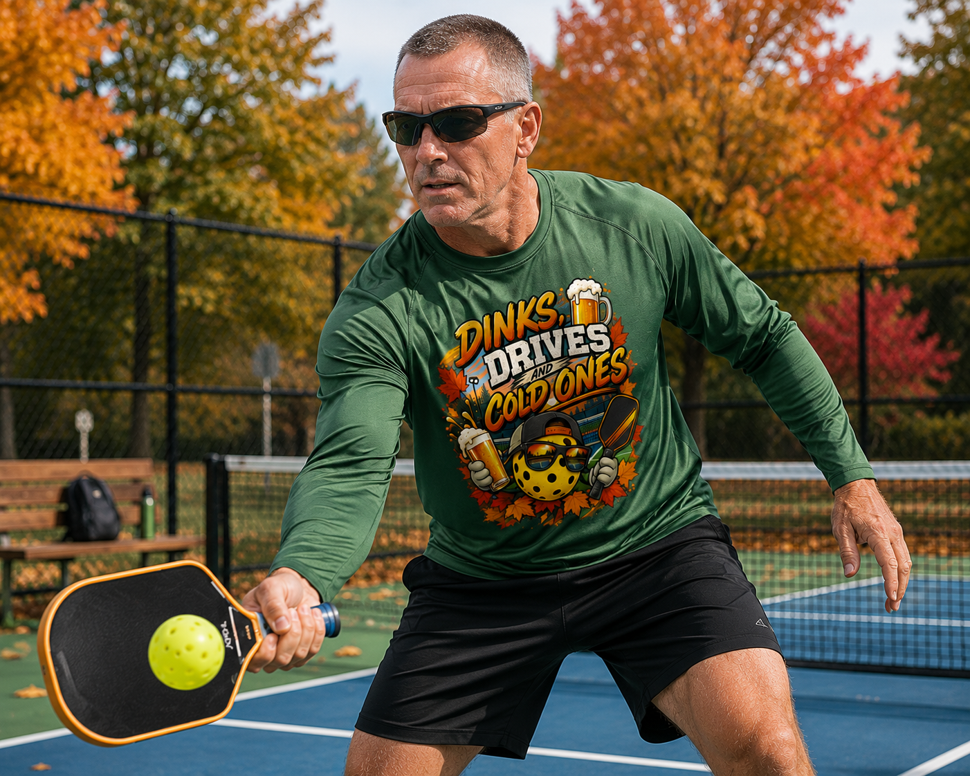 "Man wearing a green 'Dinks, Drives and Cold Ones' graphic long sleeve shirt and sunglasses while returning a low shot on an outdoor pickleball court with colorful autumn foliage in the background"