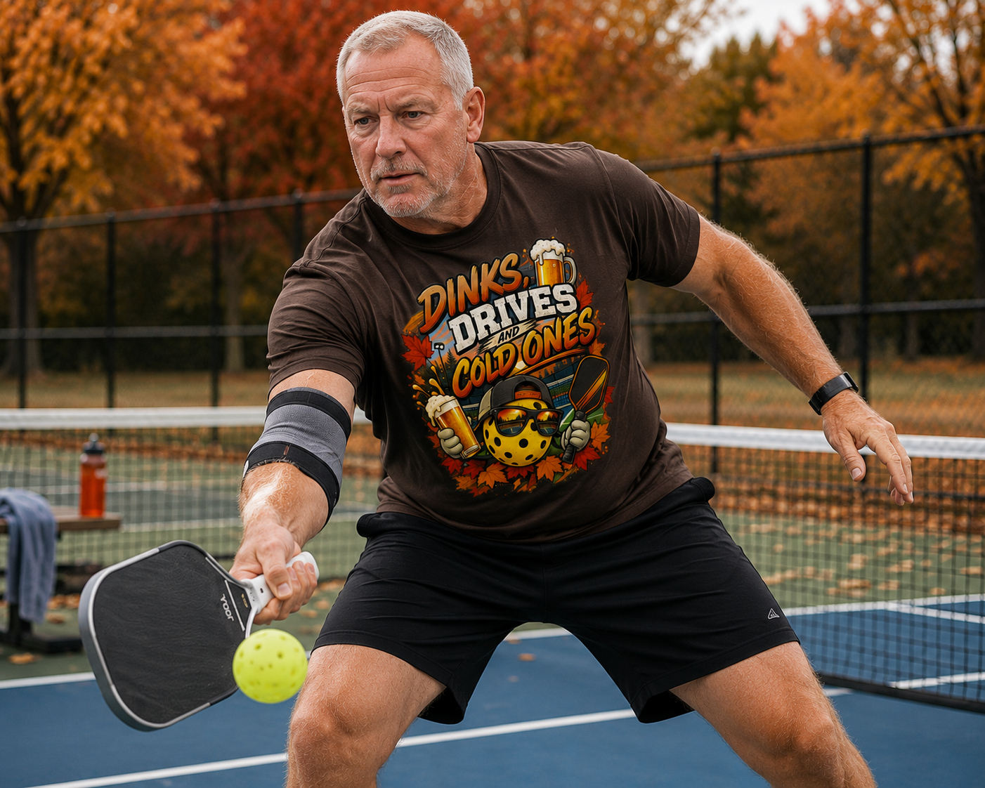 "Man wearing a brown 'Dinks, Drives and Cold Ones' graphic tee while lunging to dink at the kitchen line on an outdoor pickleball court with vibrant autumn foliage in the background"