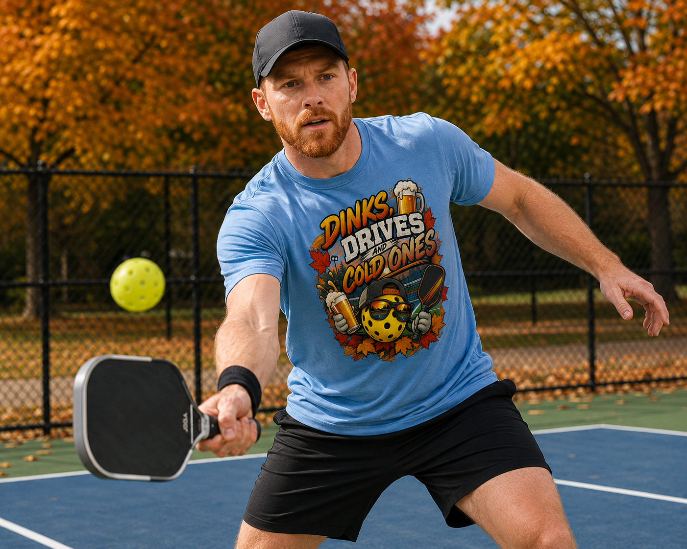 "Man wearing a blue 'Dinks, Drives and Cold Ones' graphic tee and black cap while reaching to return a shot on an outdoor pickleball court with autumn foliage in the background"