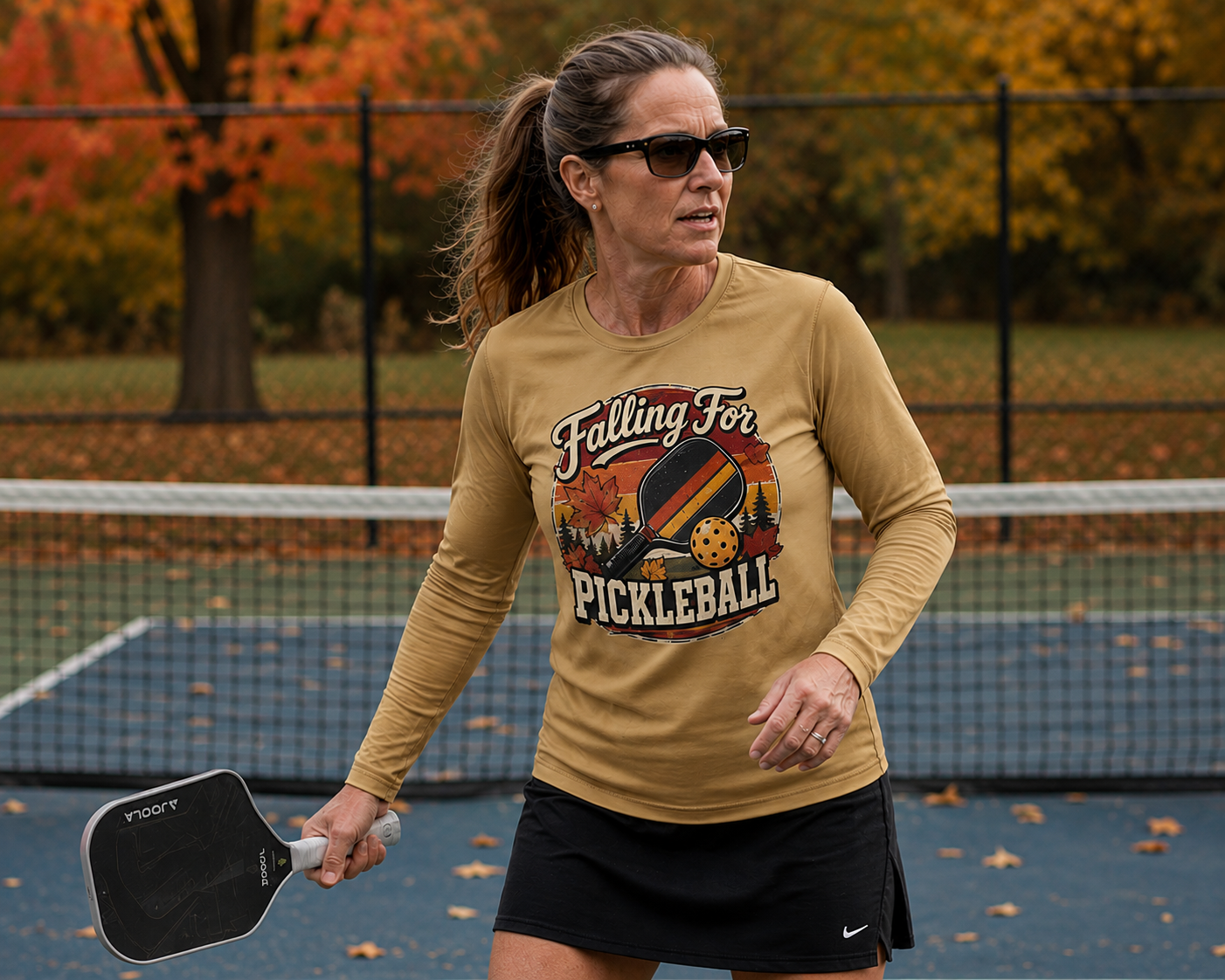 "Woman wearing a tan 'Falling For Pickleball' graphic moisture wicking long sleeve shirt and sunglasses while playing pickleball on an outdoor court surrounded by autumn foliage