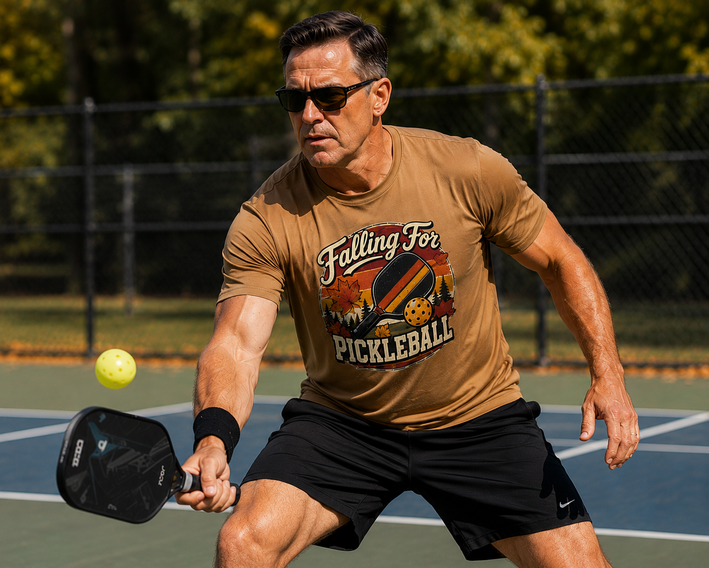 "Man wearing a tan 'Falling For Pickleball' moisture wicking graphic tee and sunglasses while lunging to return a shot on an outdoor pickleball court"