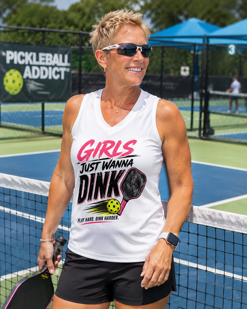 "Woman wearing white Girls Just Wanna Dink sleeveless athletic V-neck shirt on a pickleball court, holding a paddle and smiling"