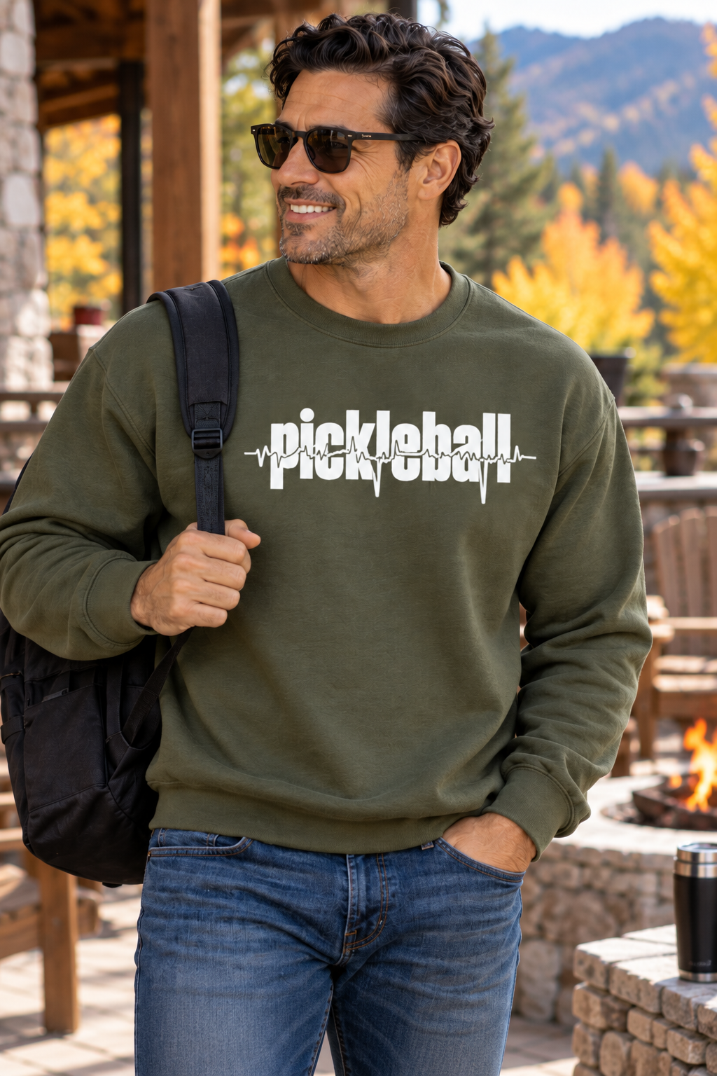 Man wearing a military green sweatshirt with white pickleball lettering and a heartbeat line, carrying a black bag outdoors with autumn trees in the background.