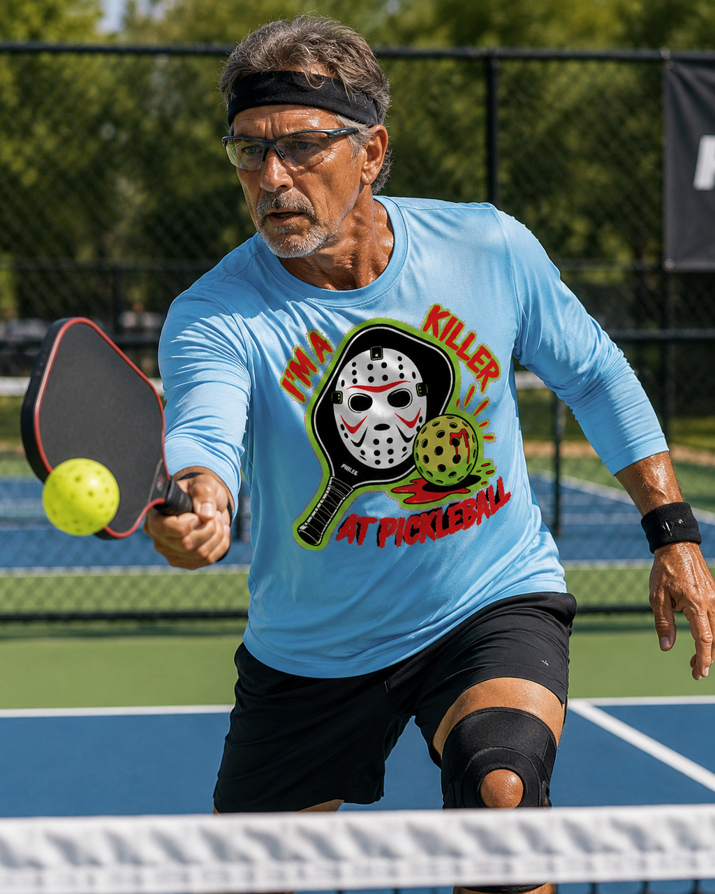 A man playing pickleball on an outdoor court, wearing a light blue long-sleeve graphic T-shirt with a horror-themed print of a hockey mask and the text 'I'M A KILLER AT PICKLEBALL.