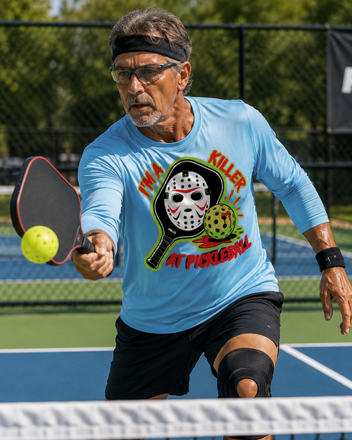 A man playing pickleball on an outdoor court, wearing a light blue long-sleeve graphic T-shirt with a horror-themed print of a hockey mask and the text 'I'M A KILLER AT PICKLEBALL.
