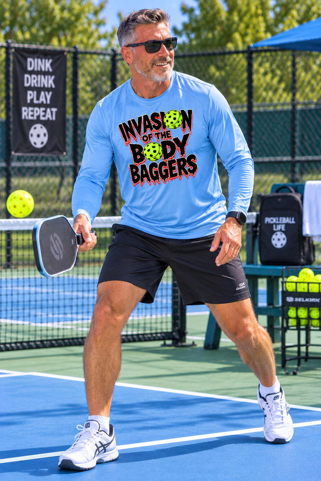 A man on a pickleball court wearing a light blue long-sleeve t-shirt with the text 'INVASION OF THE BODY BAGGERS' in red and green, paired with black shorts and white sneakers.