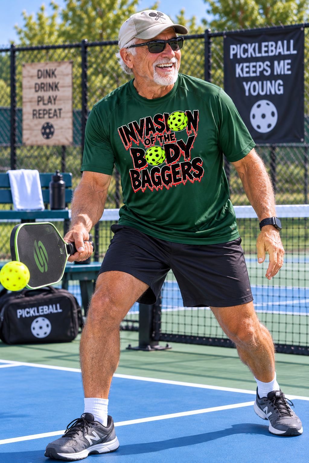 An older man playing pickleball on an outdoor court, wearing a green graphic moisture wicking shirt that reads 'INVASION OF THE BODY BAGGERS' and black shorts. He is holding a pickleball paddle and smiling, with a pickleball in the air near his hand.