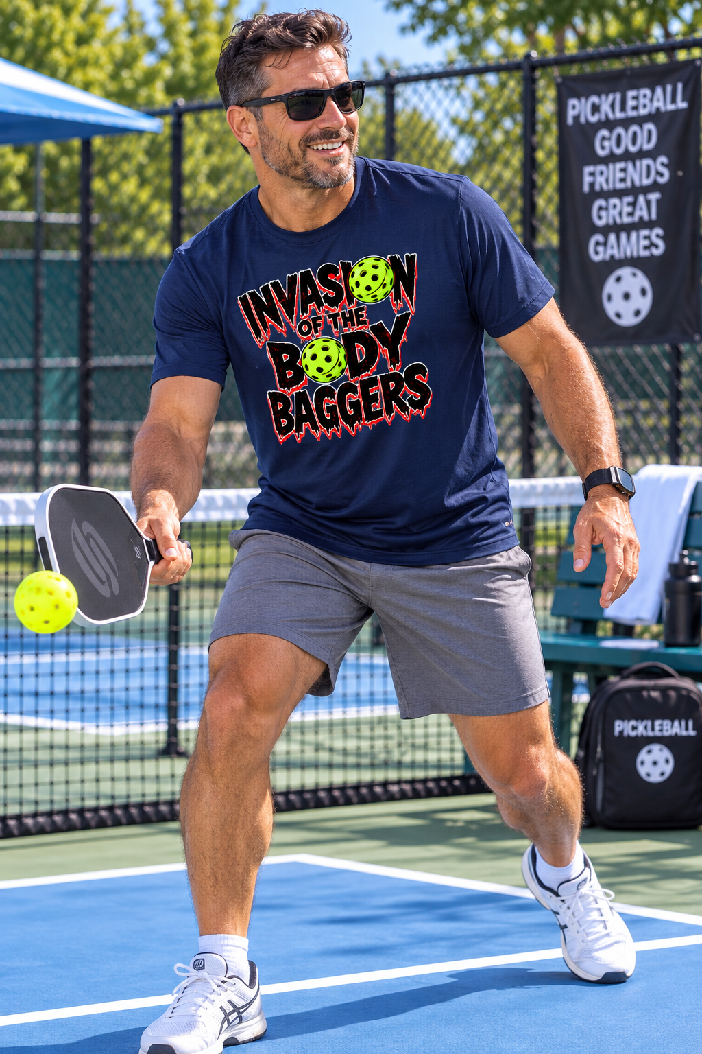 A man playing pickleball on an outdoor court wearing a navy blue graphic t-shirt that reads 'INVASION OF THE BODY BAGGERS' with green pickleballs, gray athletic shorts, and white sneakers.