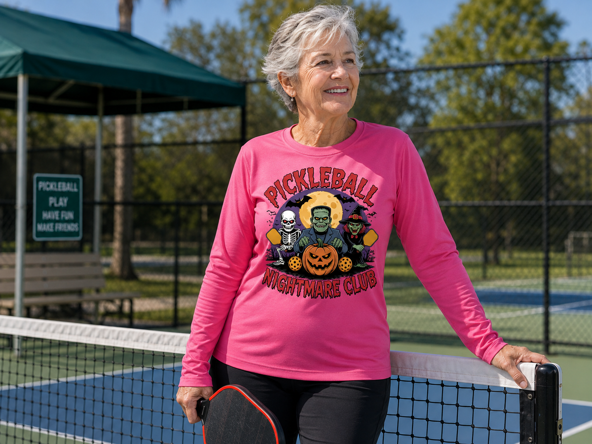 An older woman stands on an outdoor pickleball court wearing a bright pink long-sleeve graphic shirt that reads 'PICKLEBALL NIGHTMARE CLUB' with a Halloween-themed character print. She is smiling and holding a pickleball paddle, with a net, fence, trees, and a covered picnic area in the background.