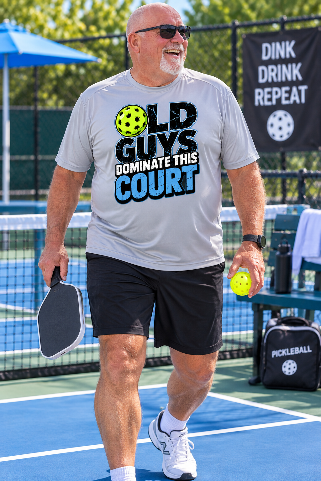 Older man holding a pickleball paddle and ball on an outdoor court while wearing a light gray “Old Guys Dominate This Court” T-shirt.