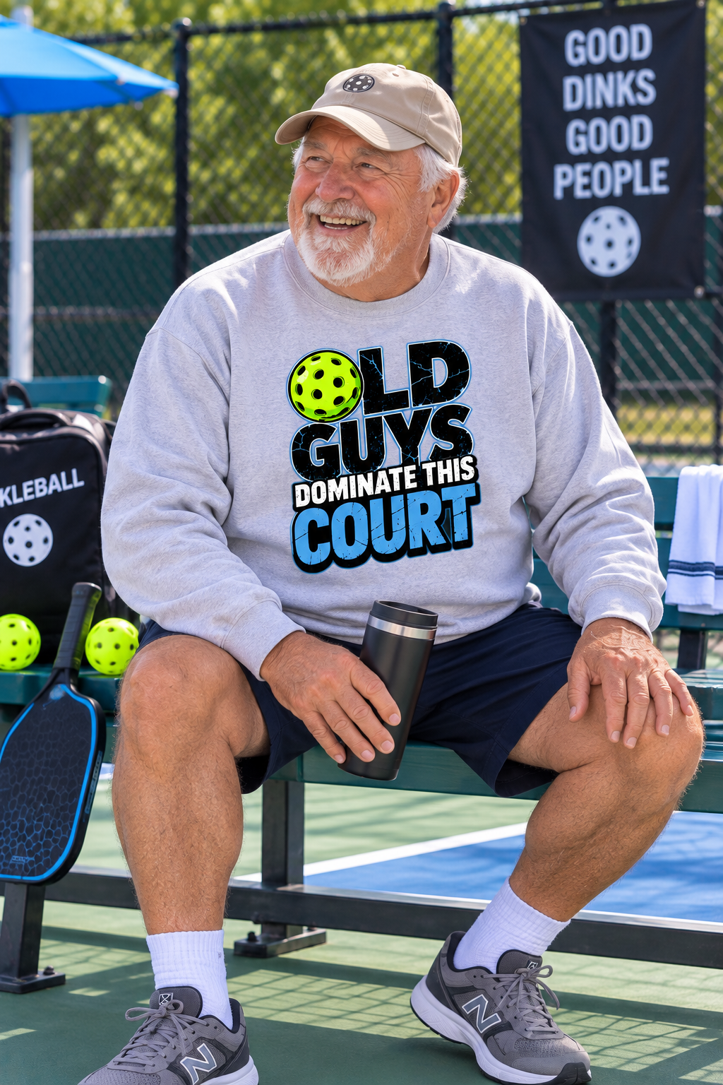 Older man wearing a light gray “Old Guys Dominate This Court” sweatshirt while sitting courtside with a pickleball paddle and tumbler.