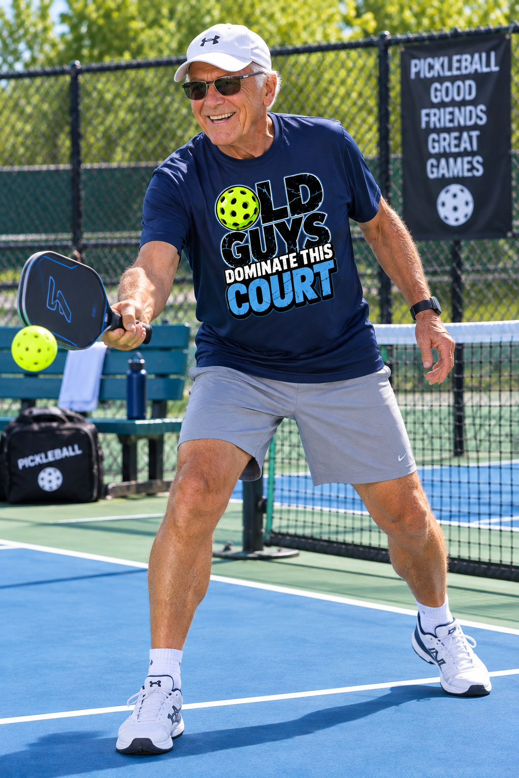 Older man playing pickleball on an outdoor court while wearing a navy “Old Guys Dominate This Court” T-shirt.