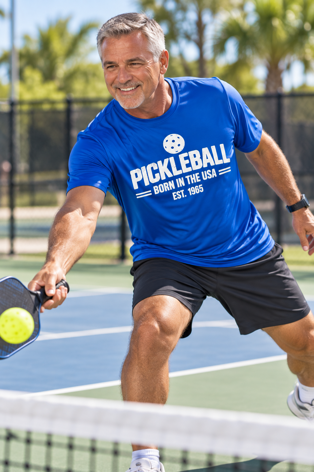 Man playing pickleball on an outdoor court wearing a blue T-shirt with white “Pickleball Born in the USA Est. 1965” lettering.