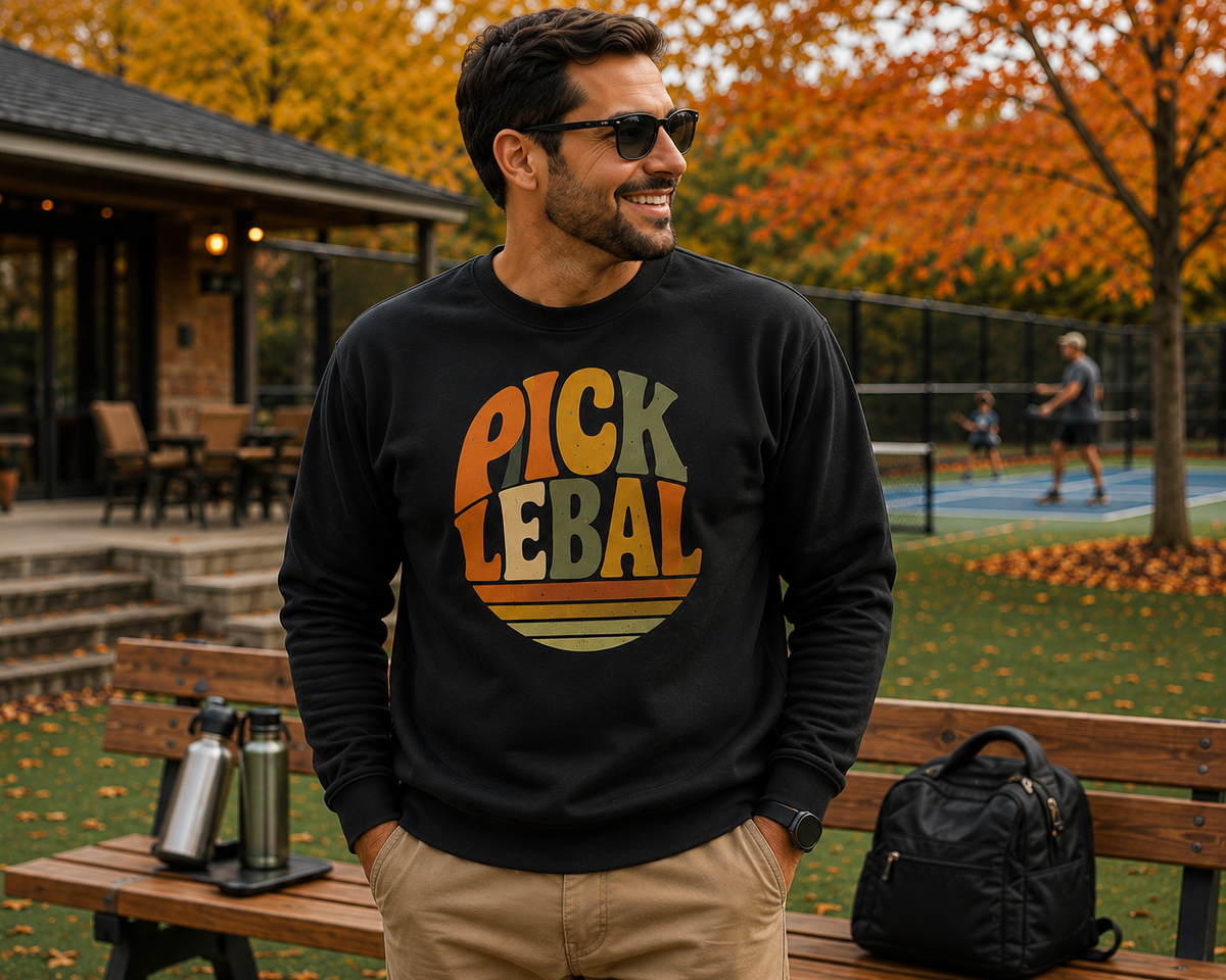 "Man smiling and wearing a black retro 'Pickleball' graphic sweatshirt and sunglasses while standing near a bench with water bottles and a bag, with players on a pickleball court and autumn foliage in the background"