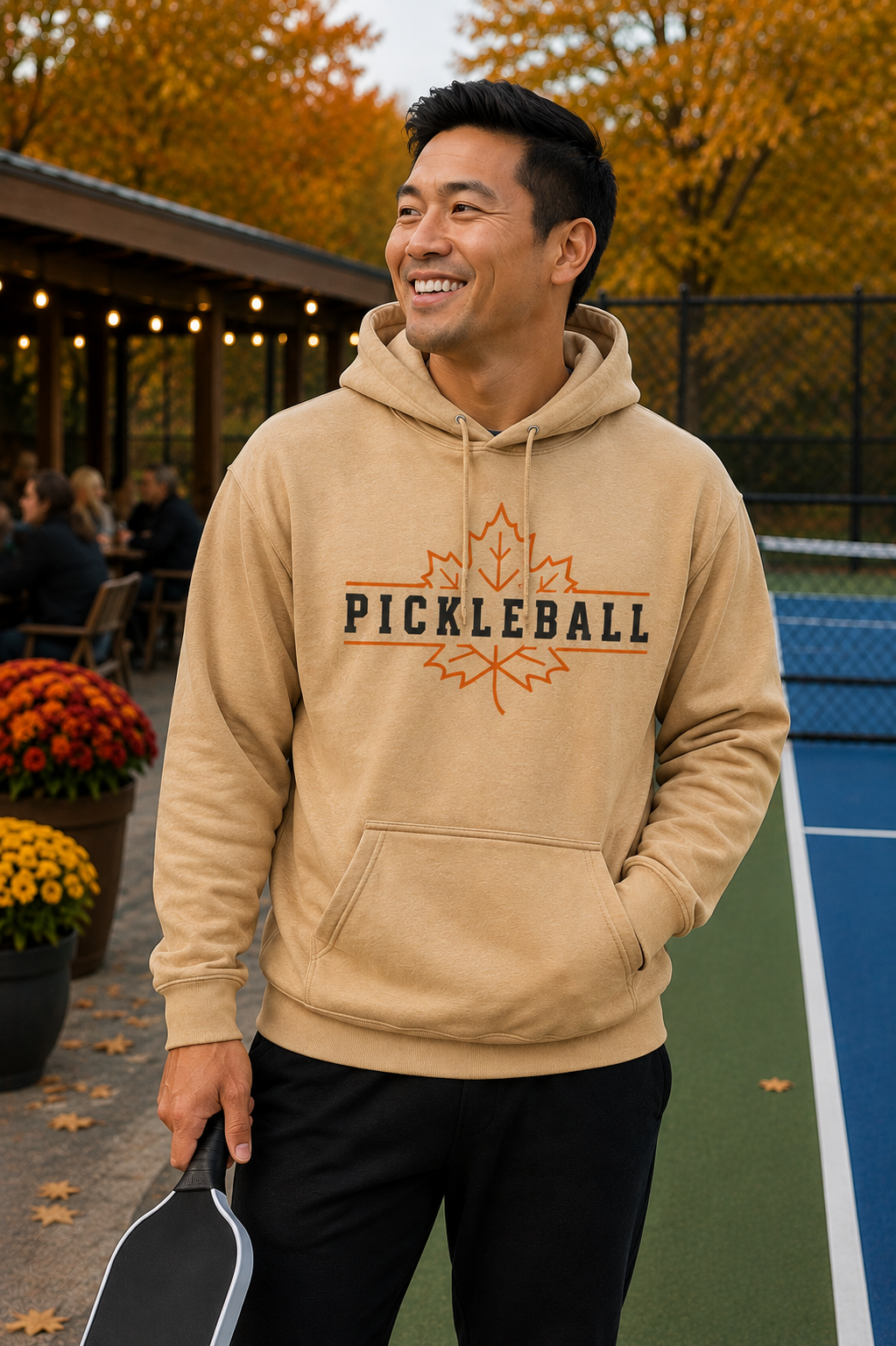 "Man smiling near an outdoor pickleball court wearing a tan 'Pickleball' maple leaf graphic pullover hoodie, holding a paddle with fall mums, string lights, and autumn foliage in the background"