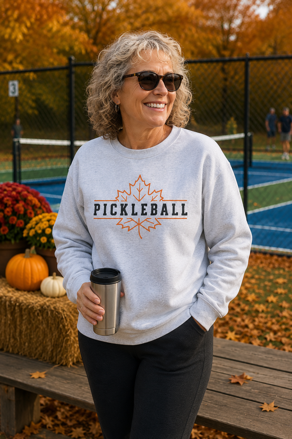 "Woman smiling near an outdoor pickleball court wearing a light gray 'Pickleball' maple leaf graphic crewneck sweatshirt, holding a travel mug with fall pumpkins, mums, hay bales, and autumn foliage in the background"