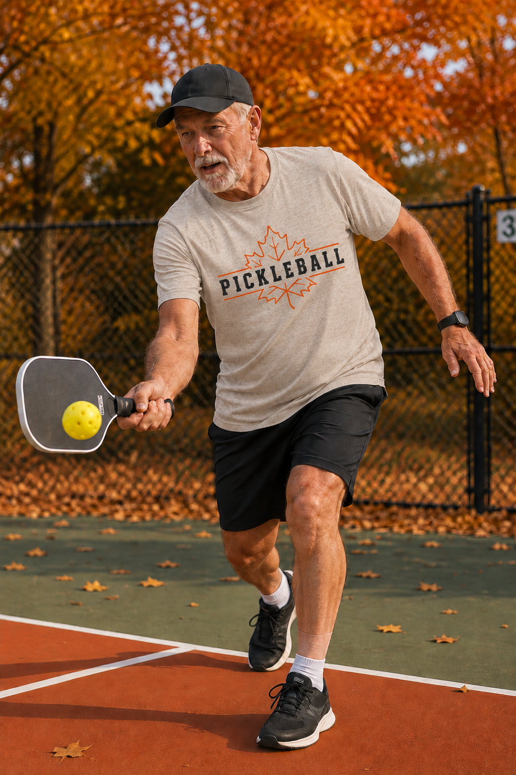 "Man actively playing pickleball on an outdoor court wearing a cream 'Pickleball' maple leaf graphic t-shirt, mid-swing with a paddle and yellow ball, vibrant fall foliage in the background"